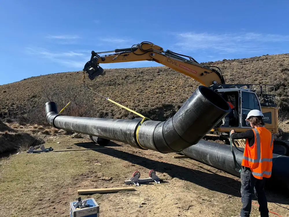 Excavator lifting a large black polyethylene pipe bend while a worker in hi-vis guides it